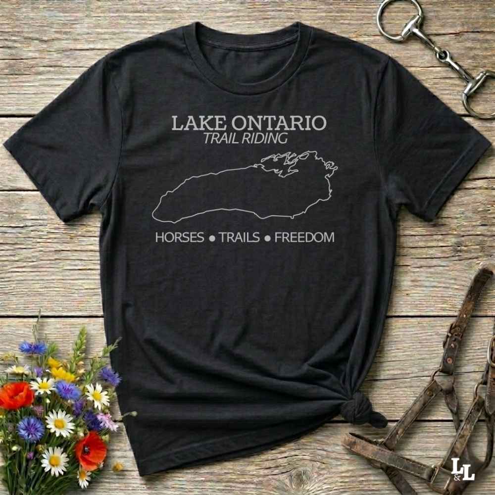 A black short-sleeve graphic T-shirt laid flat on a rustic wood surface, with white text reading 'LAKE ONTARIO TRAIL RIDING' and 'HORSES • TRAILS • FREEDOM' plus a white outline map. Decorative flowers and equestrian hardware are placed around it.
