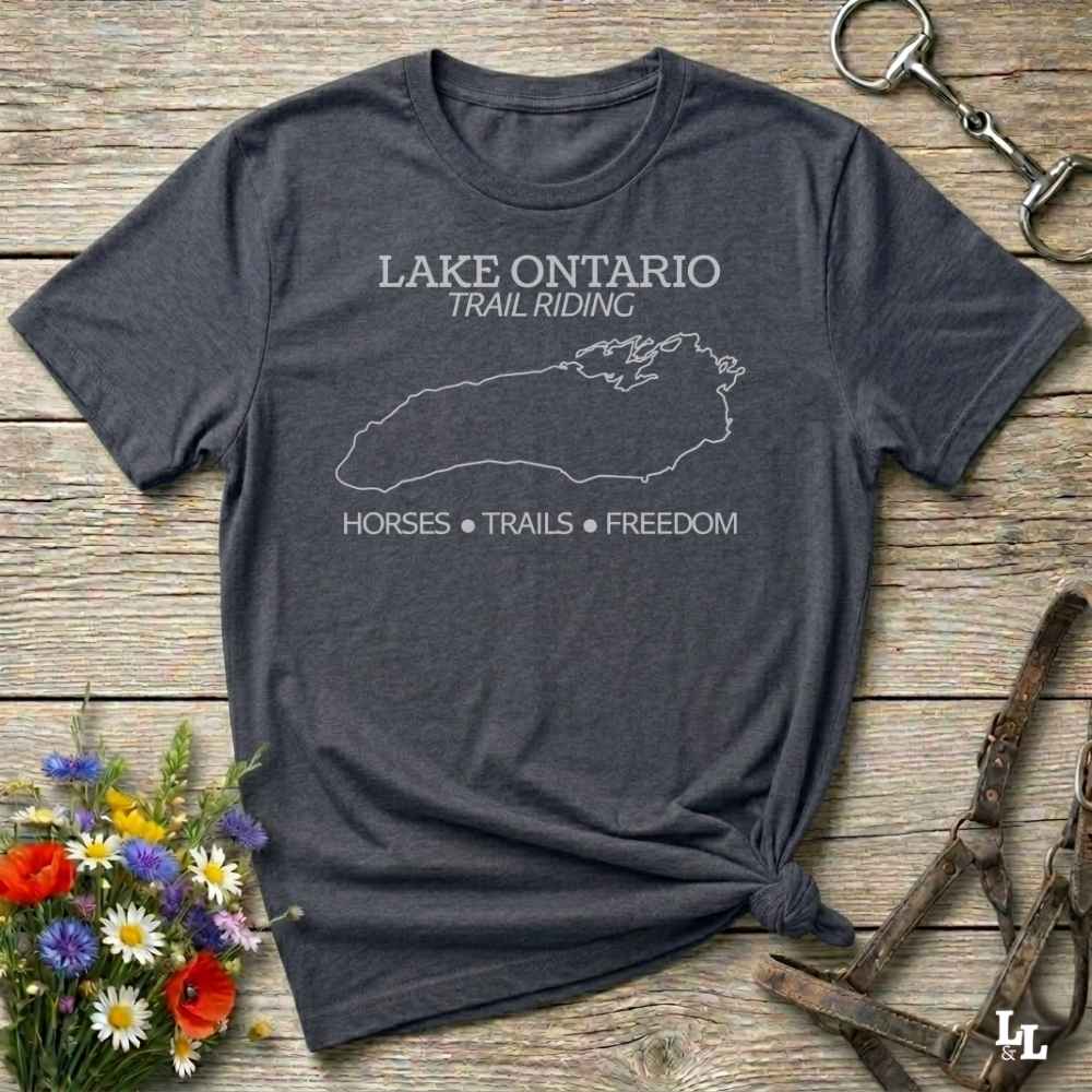 A dark heather gray graphic t-shirt laid flat on a rustic wooden surface, styled with a small bouquet of wildflowers, a horse halter, and a metal tack piece. The t-shirt features a white outline of Lake Ontario and the text 'LAKE ONTARIO TRAIL RIDING' and 'HORSES * TRAILS * FREEDOM'.