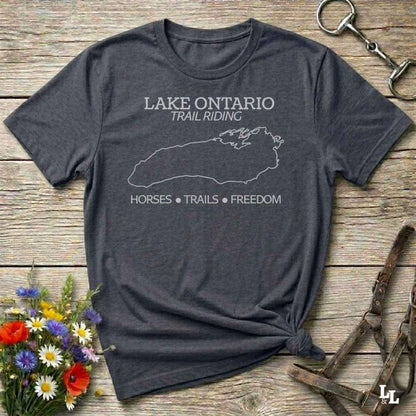A dark heather gray graphic t-shirt laid flat on a rustic wooden surface, styled with a small bouquet of wildflowers, a horse halter, and a metal tack piece. The t-shirt features a white outline of Lake Ontario and the text 'LAKE ONTARIO TRAIL RIDING' and 'HORSES * TRAILS * FREEDOM'.