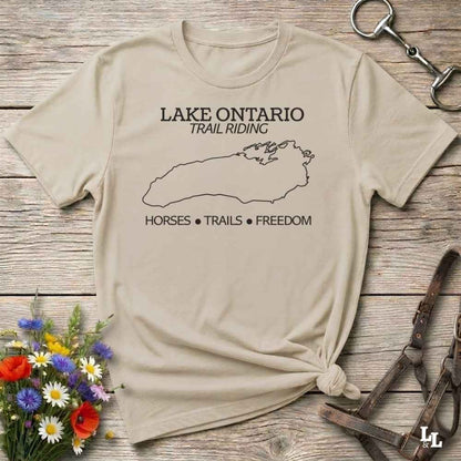 A beige graphic t-shirt laid flat on a rustic wooden surface, styled with equestrian accessories and a small flower bouquet. The shirt features a black outline of Lake Ontario and the text 'LAKE ONTARIO TRAIL RIDING' and 'HORSES • TRAILS • FREEDOM'.
