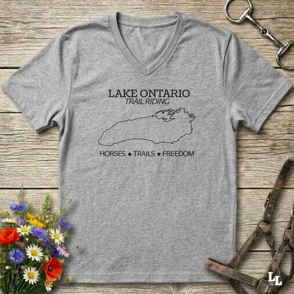 A heather gray v-neck t-shirt with a black graphic print of Lake Ontario and the text 'LAKE ONTARIO TRAIL RIDING' and 'HORSES • TRAILS • FREEDOM'. The shirt is displayed flat on a rustic wooden background with equestrian accessories and flowers.