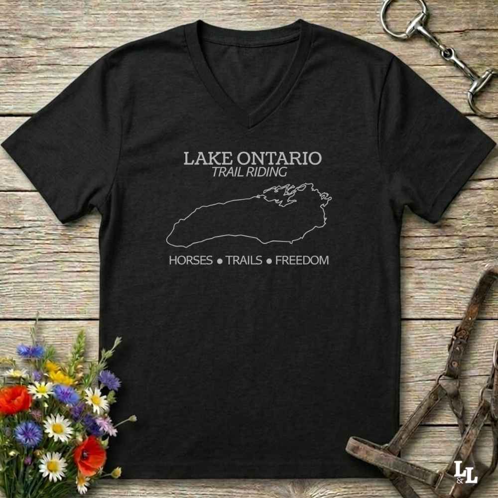 A black V-neck t-shirt with a white graphic print reading 'LAKE ONTARIO TRAIL RIDING' and 'HORSES • TRAILS • FREEDOM' around a lake outline, displayed on a rustic wooden background with equestrian accessories and flowers.