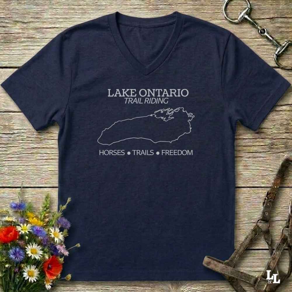 A navy blue V-neck t-shirt with a white graphic print reading 'LAKE ONTARIO TRAIL RIDING' and 'HORSES • TRAILS • FREEDOM' with a map outline, laid flat on a rustic wooden surface with horse tack and flowers as props.