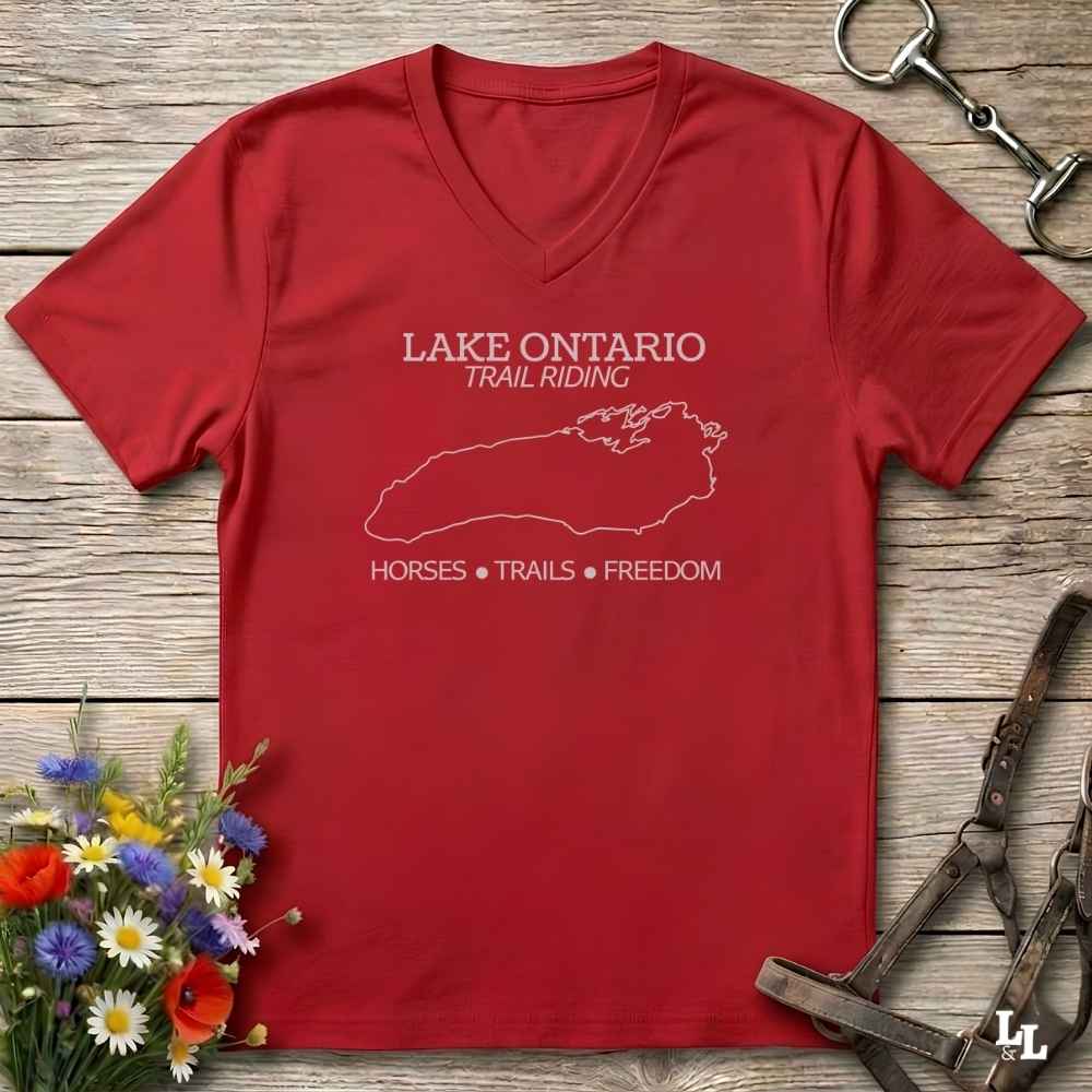 A red V-neck t-shirt with a white graphic print reading 'LAKE ONTARIO TRAIL RIDING' and 'HORSES • TRAILS • FREEDOM' with a map outline, laid flat on a rustic wood background with equestrian accessories and flowers.