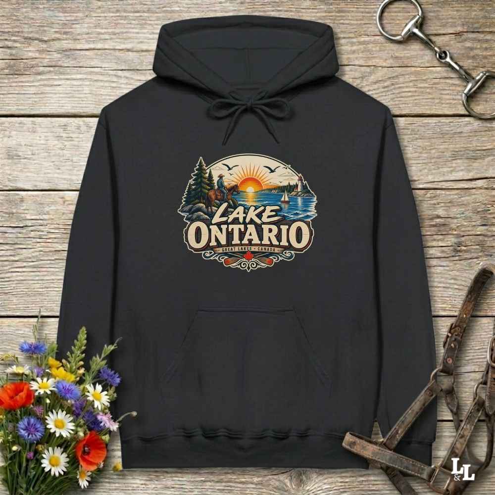 A black pullover hoodie laid flat on a rustic wooden surface, featuring a colorful retro-style 'Lake Ontario' graphic on the chest. The design includes a sunset over water, pine trees, and a lake scene.