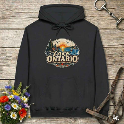 A black pullover hoodie laid flat on a rustic wooden surface, featuring a colorful retro-style 'Lake Ontario' graphic on the chest. The design includes a sunset over water, pine trees, and a lake scene.