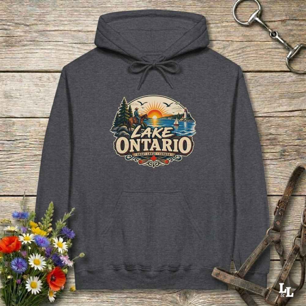 A dark heather gray hoodie laid flat on a rustic wooden surface, featuring a colorful 'Lake Ontario' scenic graphic on the chest. The hoodie is styled with flowers on one side and horse tack on the other.