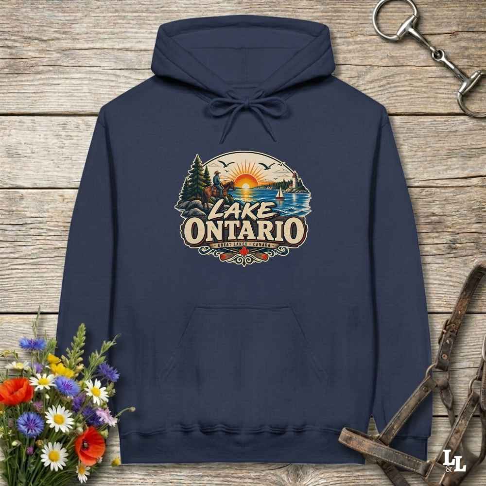 A navy blue pullover hoodie with a colorful 'Lake Ontario' graphic on the chest, displayed flat on a rustic wooden background with decorative flowers and horse tack.