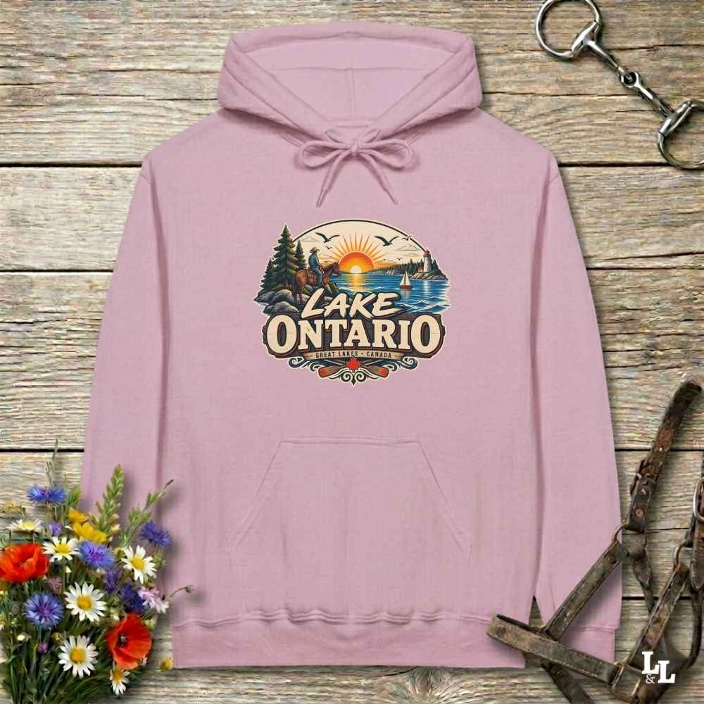 A dusty pink pullover hoodie laid flat on a rustic wooden surface, featuring a colorful retro-style 'Lake Ontario' graphic on the chest. The hoodie is styled with a bouquet of wildflowers on the bottom left, a metal horse bit in the top right, and leather reins at the bottom right.