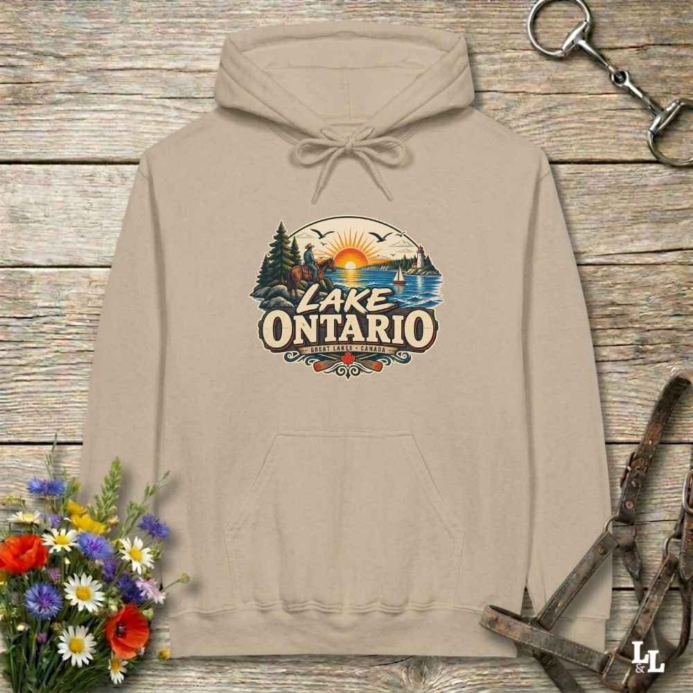 A beige pullover hoodie laid flat on a rustic wood plank surface, featuring a colorful 'Lake Ontario' scenic graphic on the chest. The hoodie is styled with a small bouquet of flowers at the lower left, a metal tack item at the upper right, and a leather strap at the lower right.