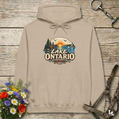 A beige pullover hoodie laid flat on a rustic wood plank surface, featuring a colorful 'Lake Ontario' scenic graphic on the chest. The hoodie is styled with a small bouquet of flowers at the lower left, a metal tack item at the upper right, and a leather strap at the lower right.