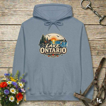 A slate blue pullover hoodie with a 'Lake Ontario' scenic graphic on the chest, laid flat on a rustic wooden background with flowers and horse tack props.