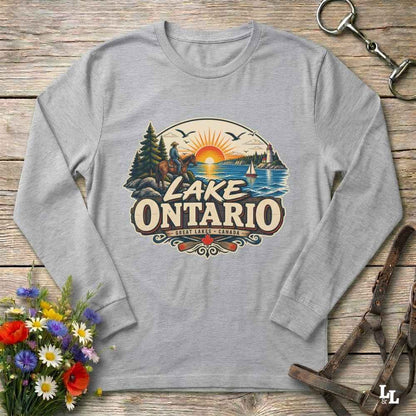 A light gray long-sleeve t-shirt with a vintage-style 'Lake Ontario' graphic, laid flat on a rustic wooden surface with decorative flowers and leather tack around it.