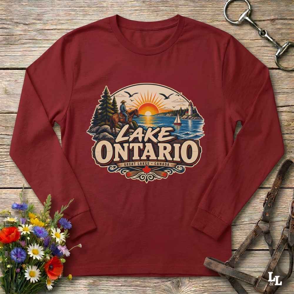 A long-sleeve crewneck shirt in maroon red, laid flat on a rustic wooden surface. The shirt features a large graphic print on the chest depicting a scenic sunset over Lake Ontario with a horseback rider, pine trees, and a sailboat.