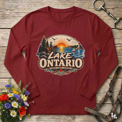 A long-sleeve crewneck shirt in maroon red, laid flat on a rustic wooden surface. The shirt features a large graphic print on the chest depicting a scenic sunset over Lake Ontario with a horseback rider, pine trees, and a sailboat.