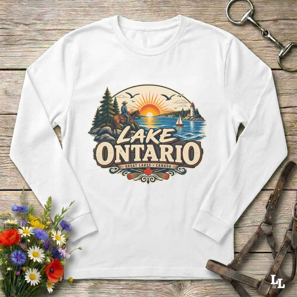 A white long-sleeve crewneck t-shirt with a colorful 'Lake Ontario' graphic print, laid flat on a rustic wooden background with decorative flowers and leather gear.