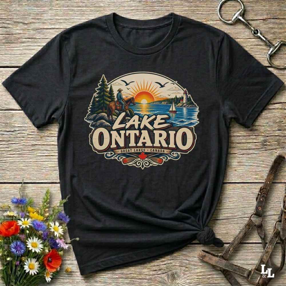 A black short-sleeve graphic T-shirt laid flat on a rustic wooden surface, featuring a colorful Lake Ontario sunset illustration with the words 'LAKE ONTARIO' and 'Great Lakes' Decorative flowers and equestrian tack are placed around the shirt.