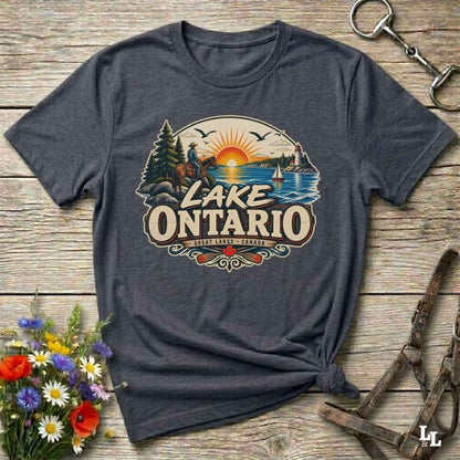 A dark heather gray graphic t-shirt featuring a retro-style 'Lake Ontario' sunset illustration, styled in a flat-lay on a rustic wooden background with a horse halter and wildflowers.