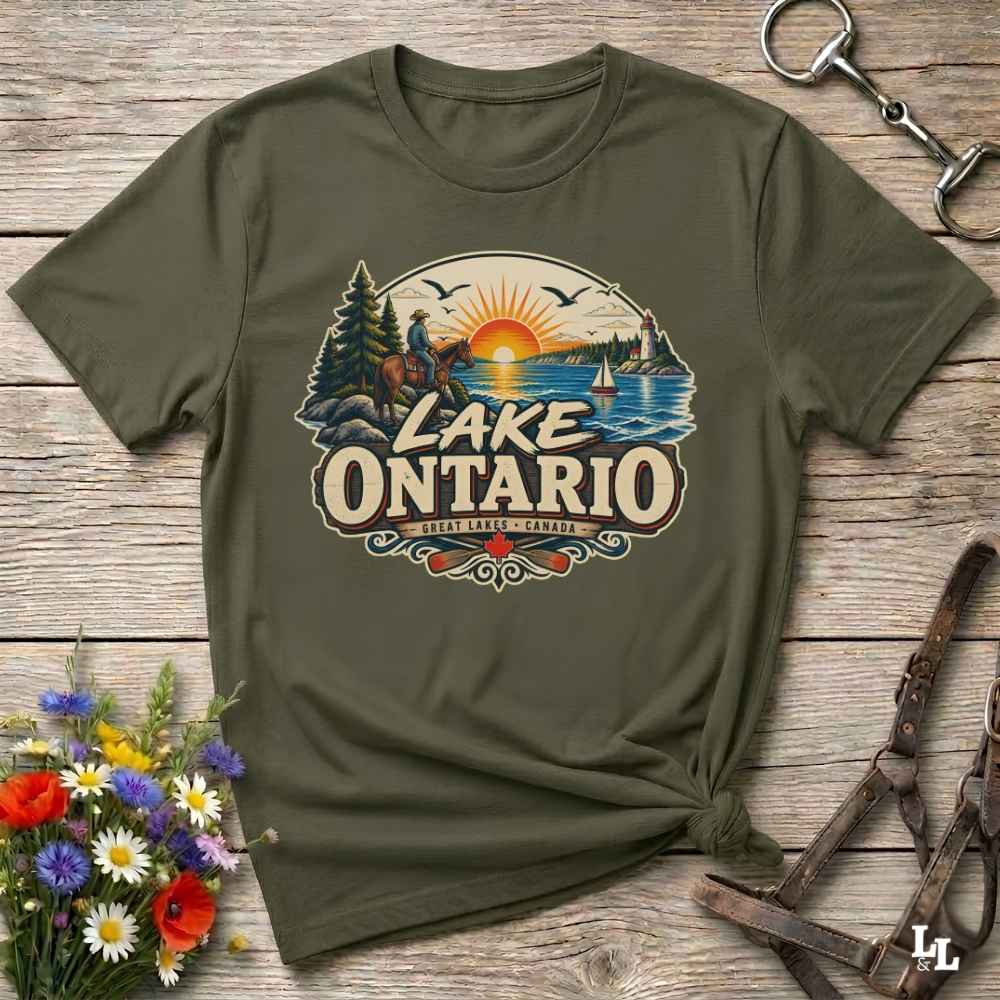 An olive green short-sleeve t-shirt with a vintage-style 'Lake Ontario' graphic print, styled in a flat-lay on a rustic wooden background with a horse bit and flowers.