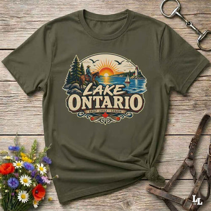 An olive green short-sleeve t-shirt with a vintage-style 'Lake Ontario' graphic print, styled in a flat-lay on a rustic wooden background with a horse bit and flowers.