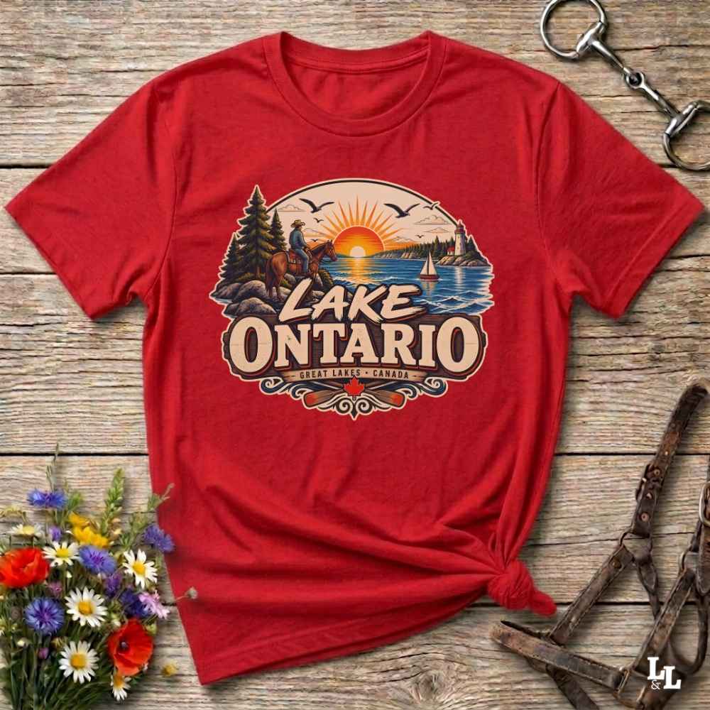 A red short-sleeve graphic T-shirt laid flat on a rustic wooden surface, with a colorful Lake Ontario sunset print on the chest. The shirt is styled with flowers, a metal ring, and leather straps around it.