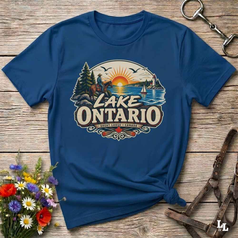 A navy blue short-sleeve t-shirt with a 'Lake Ontario' graphic print, styled in a flat-lay on a rustic wooden background with flowers and leather tack.