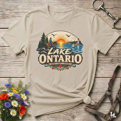 A beige graphic t-shirt featuring a retro-style 'Lake Ontario' sunset illustration, styled in a flat-lay on a rustic wooden background with flowers and leather accessories.
