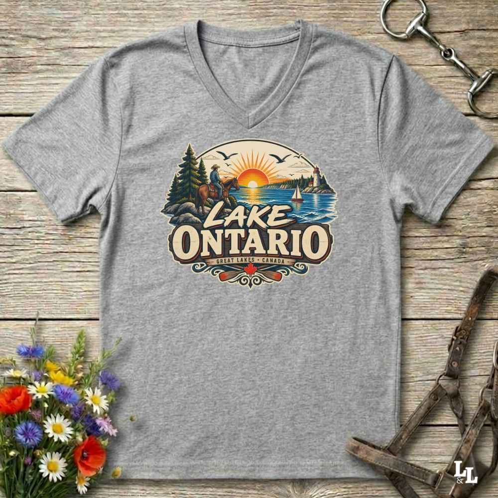 A heather gray V-neck t-shirt laid flat on a rustic wooden surface, featuring a colorful graphic of a horse rider, a sunset over Lake Ontario, and the text 'LAKE ONTARIO' with 'SURREY, ONTARIO' below. Decorative flowers and horse tack are placed around the shirt.