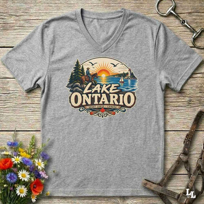 A heather gray V-neck t-shirt laid flat on a rustic wooden surface, featuring a colorful graphic of a horse rider, a sunset over Lake Ontario, and the text 'LAKE ONTARIO' with 'SURREY, ONTARIO' below. Decorative flowers and horse tack are placed around the shirt.