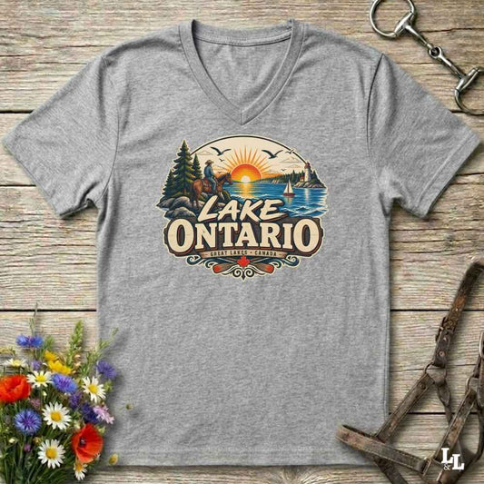 A heather gray V-neck t-shirt laid flat on a rustic wooden surface, featuring a colorful graphic of a horse rider, a sunset over Lake Ontario, and the text 'LAKE ONTARIO' with 'SURREY, ONTARIO' below. Decorative flowers and horse tack are placed around the shirt.