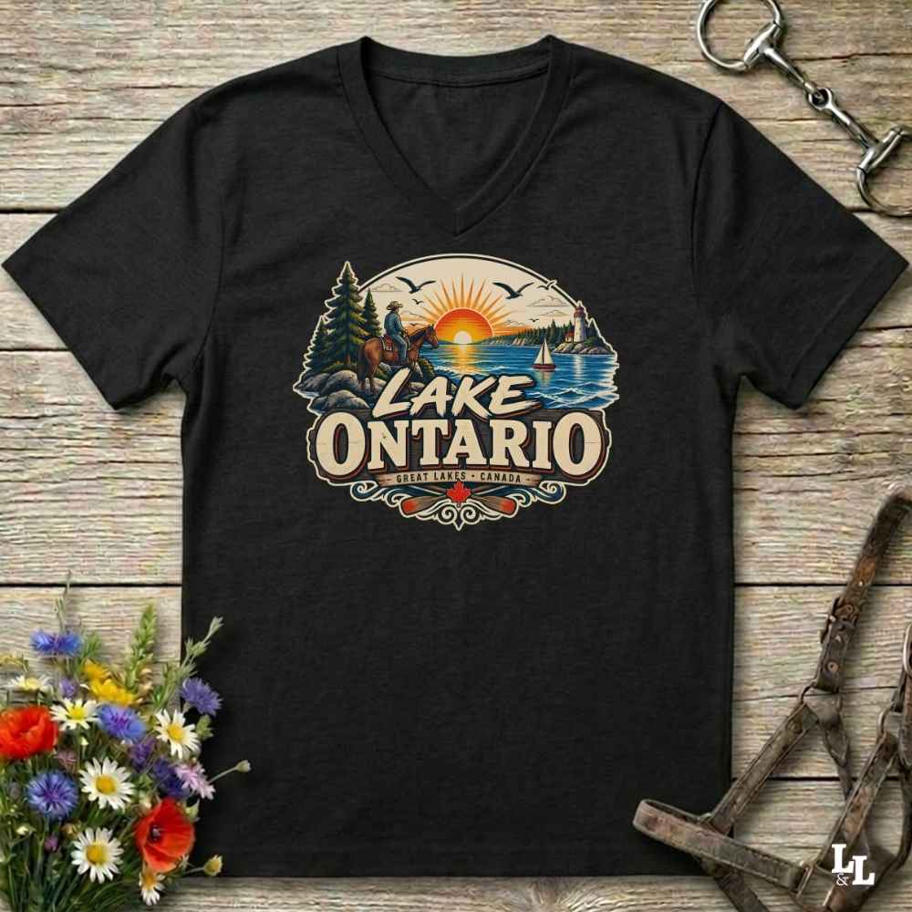 A black V-neck t-shirt with a colorful retro-style 'Lake Ontario' graphic, displayed flat on a rustic wooden background with equestrian and floral props.