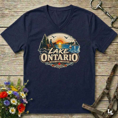 A navy blue V-neck t-shirt laid flat on a rustic wooden surface, featuring a colorful retro-style graphic of Lake Ontario with a sunset, trees, and the text 'LAKE ONTARIO'. The shirt is styled with equestrian gear and a bouquet of wildflowers.
