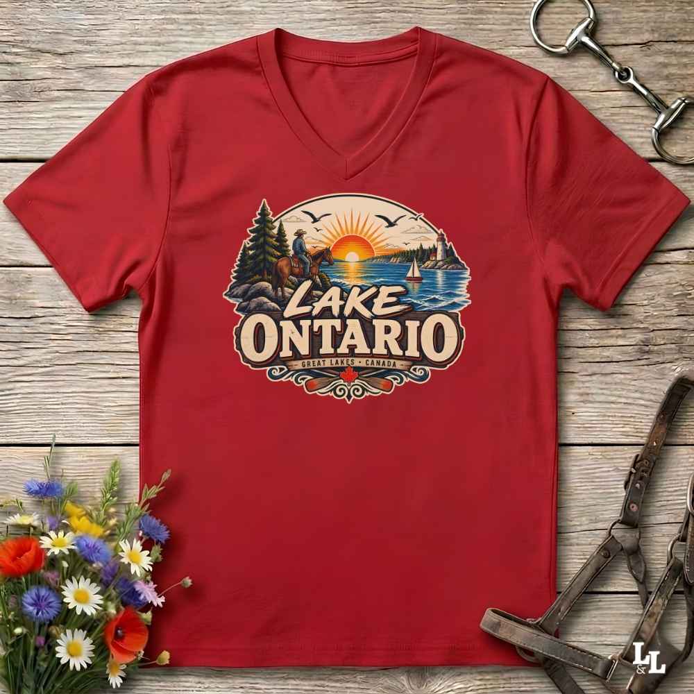 A red V-neck t-shirt laid flat on a rustic wooden surface, featuring a large circular graphic of a lake landscape with the text 'LAKE ONTARIO' and 'Great Lakes Ontario'. The shirt is styled with flowers on the bottom left and equestrian tack on the top right.