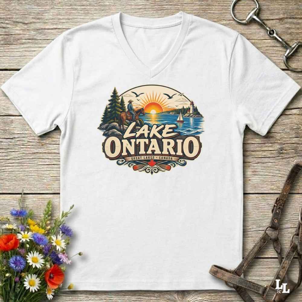 A white V-neck t-shirt laid flat on a rustic wooden surface, featuring a colorful retro-style graphic of a lake sunset with trees, a lighthouse, and the text 'LAKE ONTARIO' and 'Great Lakes' Decorative flowers and leather straps are placed around the shirt.