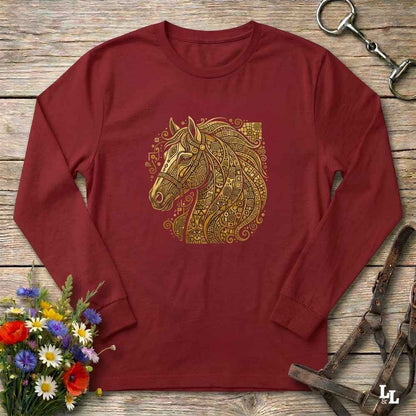 A long-sleeve crewneck t-shirt in maroon red, featuring a detailed gold and yellow horse head graphic on the chest. The shirt is styled in a flat-lay arrangement on a rustic wooden background, surrounded by equestrian tack (reins and bridle) and a small bouquet of colorful wildflowers.