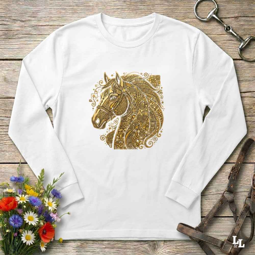 A white long-sleeve t-shirt with a detailed gold horse head graphic on the chest, laid flat on a rustic wooden surface with equestrian accessories and flowers.
