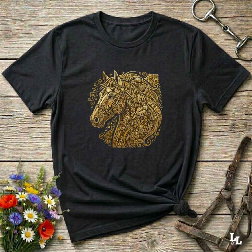 A black short-sleeve t-shirt with a detailed gold horse head graphic on the chest, styled in a flat-lay on a rustic wooden background with equestrian accessories and flowers.