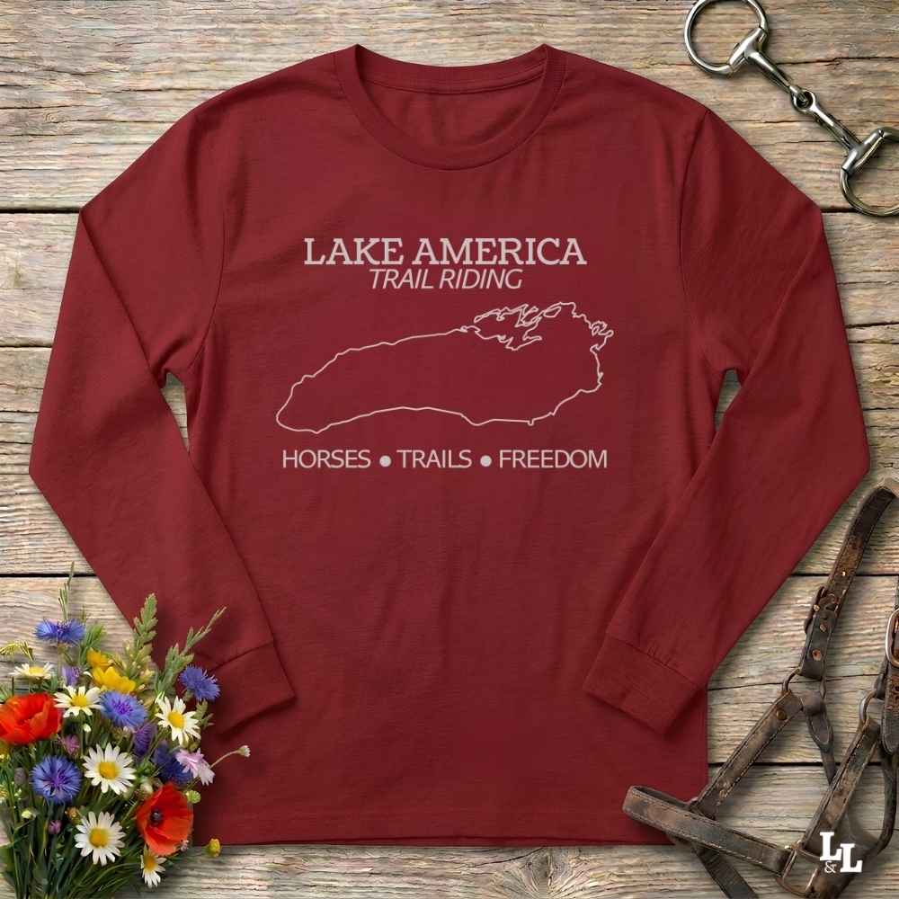 A long-sleeve crewneck t-shirt in maroon red, laid flat on a rustic wooden surface. The shirt features a white graphic print with the text 'LAKE AMERICA TRAIL RIDING' and 'HORSES • TRAILS • FREEDOM' surrounding a map outline of Lake Michigan.