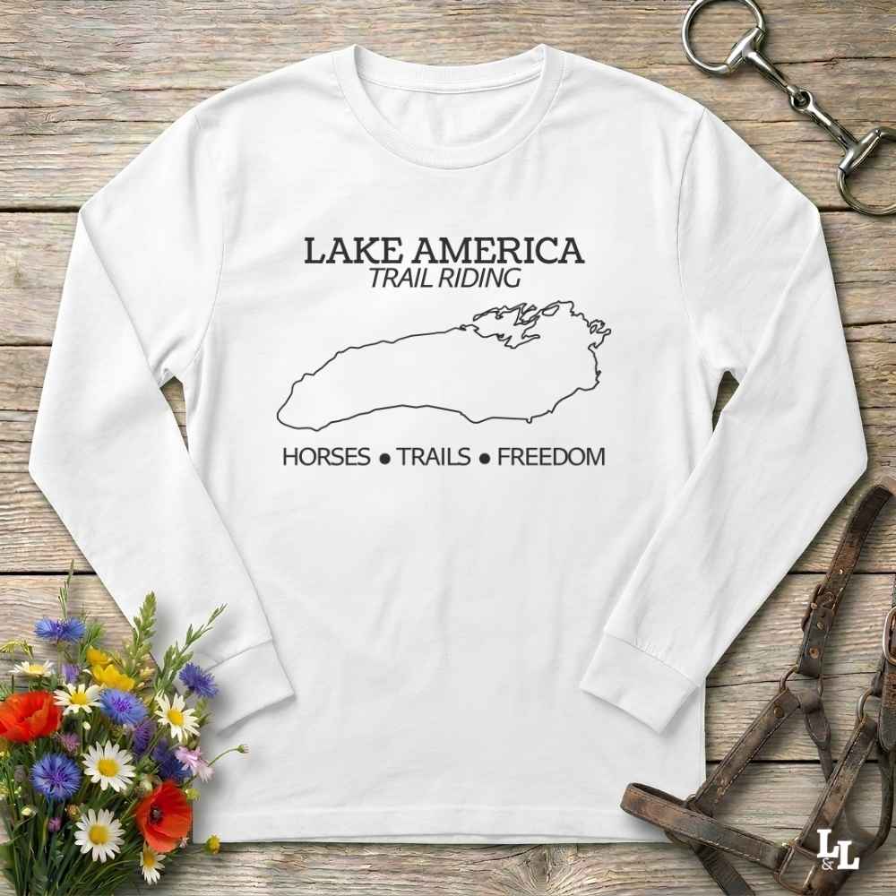 A white long-sleeve t-shirt with a white graphic print reading 'LAKE AMERICA TRAIL RIDING' and 'HORSES • TRAILS • FREEDOM' with a lake outline, laid flat on a rustic wood background with flowers and horse tack props.