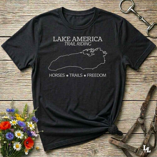 A black short-sleeve graphic T-shirt laid flat on a rustic wood surface, with white text reading 'LAKE AMERICA TRAIL RIDING' and 'HORSES • TRAILS • FREEDOM' plus a white outline of Lake Superior. The shirt is styled with flowers, a horse bit, and leather tack around it.