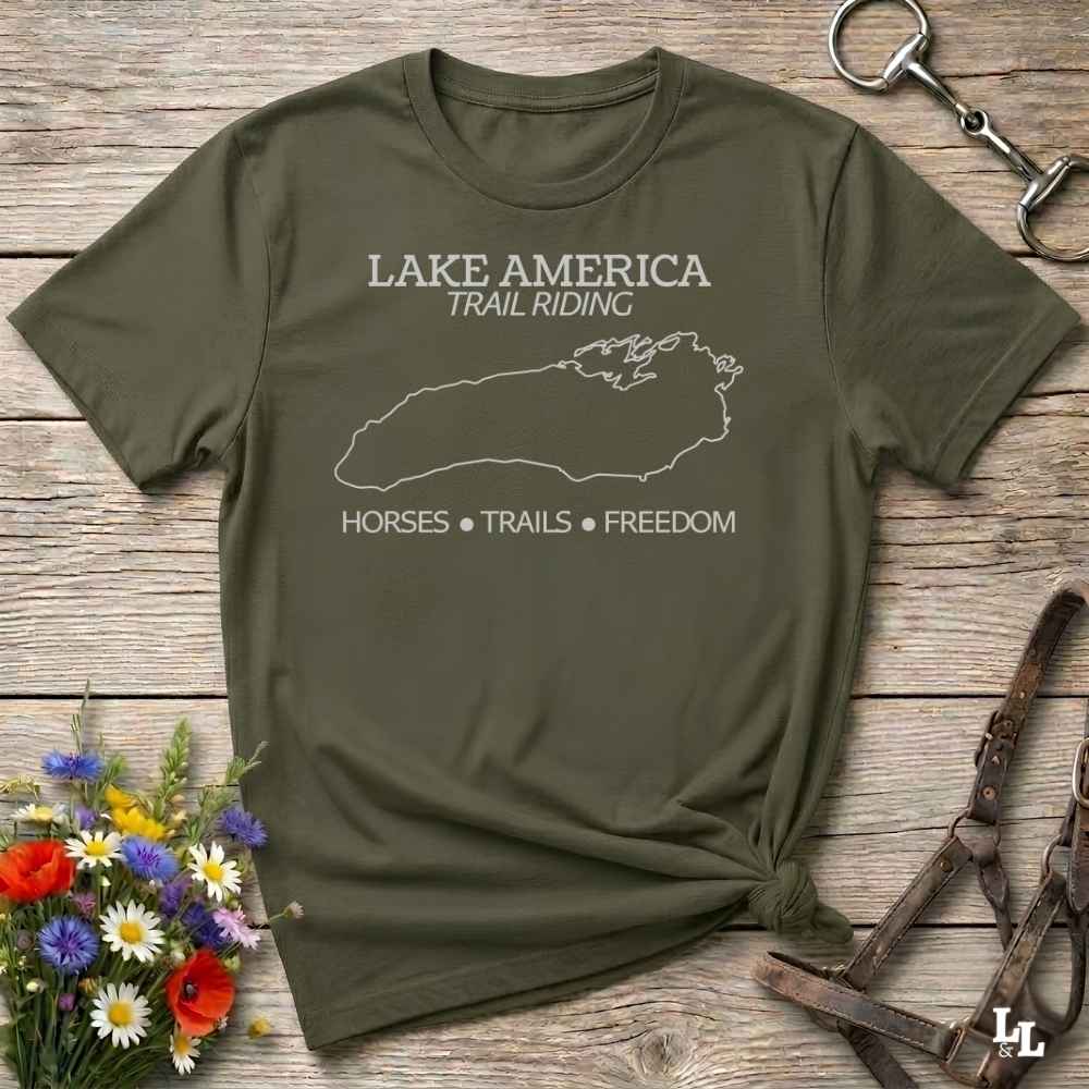 An olive green graphic t-shirt laid flat on a rustic wooden surface, styled with equestrian accessories and a small bouquet of wildflowers. The t-shirt features a white outline of Lake Superior and the text 'LAKE AMERICA TRAIL RIDING' and 'HORSES • TRAILS • FREEDOM'.