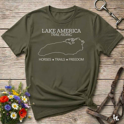 An olive green graphic t-shirt laid flat on a rustic wooden surface, styled with equestrian accessories and a small bouquet of wildflowers. The t-shirt features a white outline of Lake Superior and the text 'LAKE AMERICA TRAIL RIDING' and 'HORSES • TRAILS • FREEDOM'.