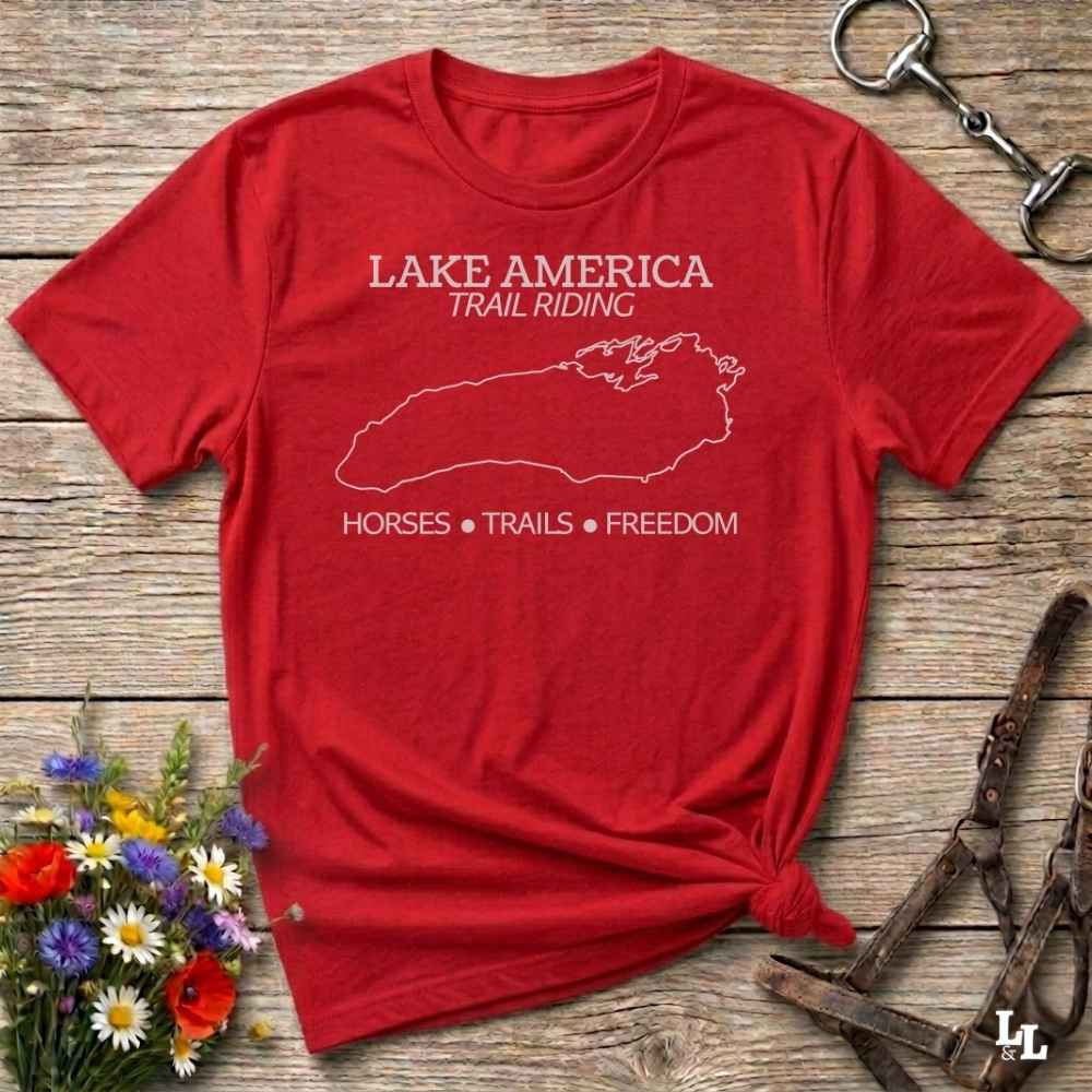 A red short-sleeve graphic T-shirt laid flat on a rustic wooden surface, with white text reading 'LAKE AMERICA TRAIL RIDING' and 'HORSES • TRAILS • FREEDOM' plus a white outline map. Decorative flowers and equestrian tack are placed around it, but no person or real environment is the main subject, so this is Product/Packaging rather than Lifestyle.