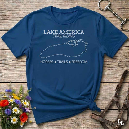 A navy blue graphic t-shirt laid flat on a rustic wooden surface, styled with equestrian accessories and wildflowers. The shirt features a white outline of Lake Superior and the text 'LAKE AMERICA TRAIL RIDING' and 'HORSES • TRAILS • FREEDOM'.