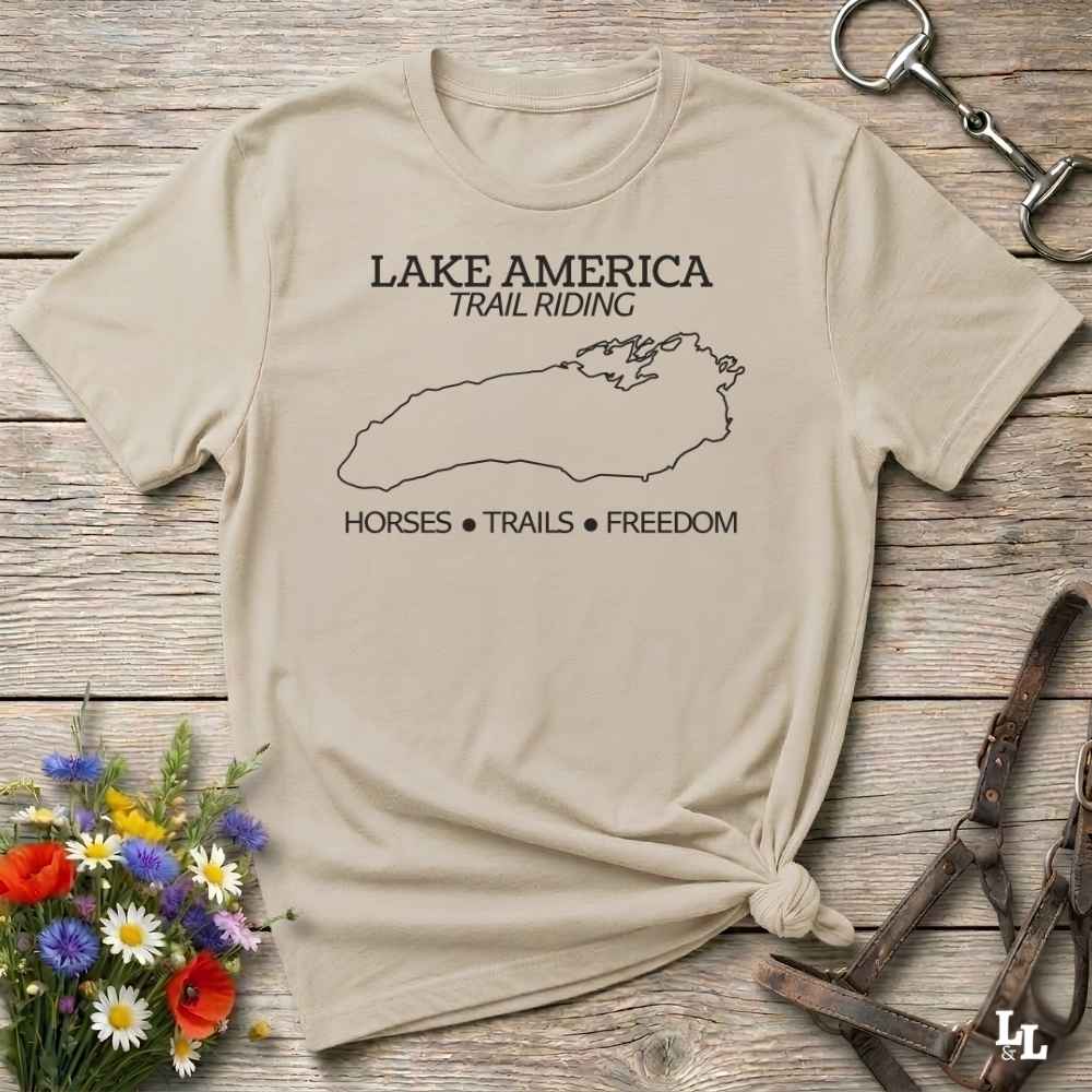 A beige graphic t-shirt laid flat on a rustic wooden surface, styled with equestrian accessories and a small flower bouquet. The shirt features a black outline map of Lake Superior with the text 'LAKE AMERICA TRAIL RIDING' and 'HORSES • TRAILS • FREEDOM'.