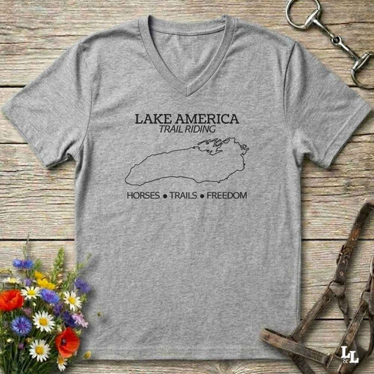 A grey  V-neck t-shirt with a white graphic print reading 'LAKE AMERICA TRAIL RIDING' and 'HORSES • TRAILS • FREEDOM' around a lake outline, displayed on a rustic wooden background with horse tack and flowers.