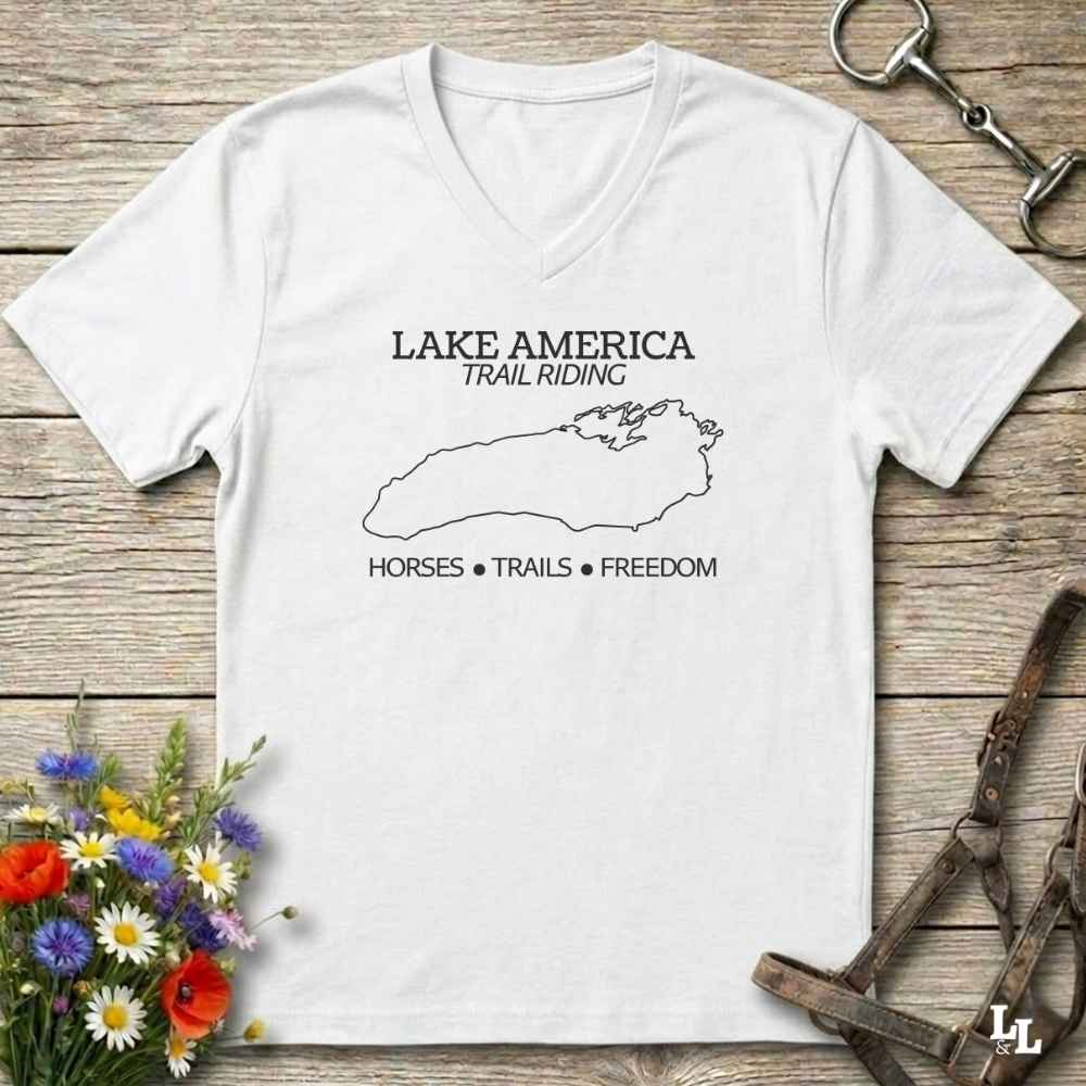 A white  V-neck t-shirt with a white graphic print reading 'LAKE AMERICA TRAIL RIDING' and 'HORSES • TRAILS • FREEDOM' around a lake outline, displayed on a rustic wooden background with horse tack and flowers.