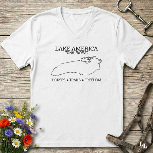 A white  V-neck t-shirt with a white graphic print reading 'LAKE AMERICA TRAIL RIDING' and 'HORSES • TRAILS • FREEDOM' around a lake outline, displayed on a rustic wooden background with horse tack and flowers.