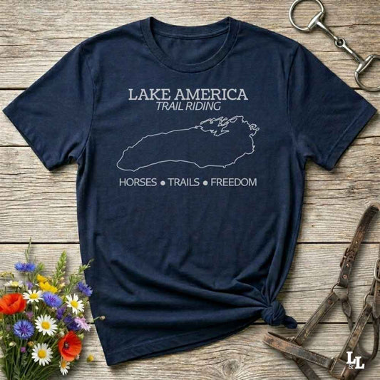A navy blue graphic t-shirt with a white outline of Lake Superior and the text 'LAKE AMERICA TRAIL RIDING' and 'HORSES * TRAILS * FREEDOM', styled in a flat lay on a rustic wooden background with equestrian accessories and flowers.