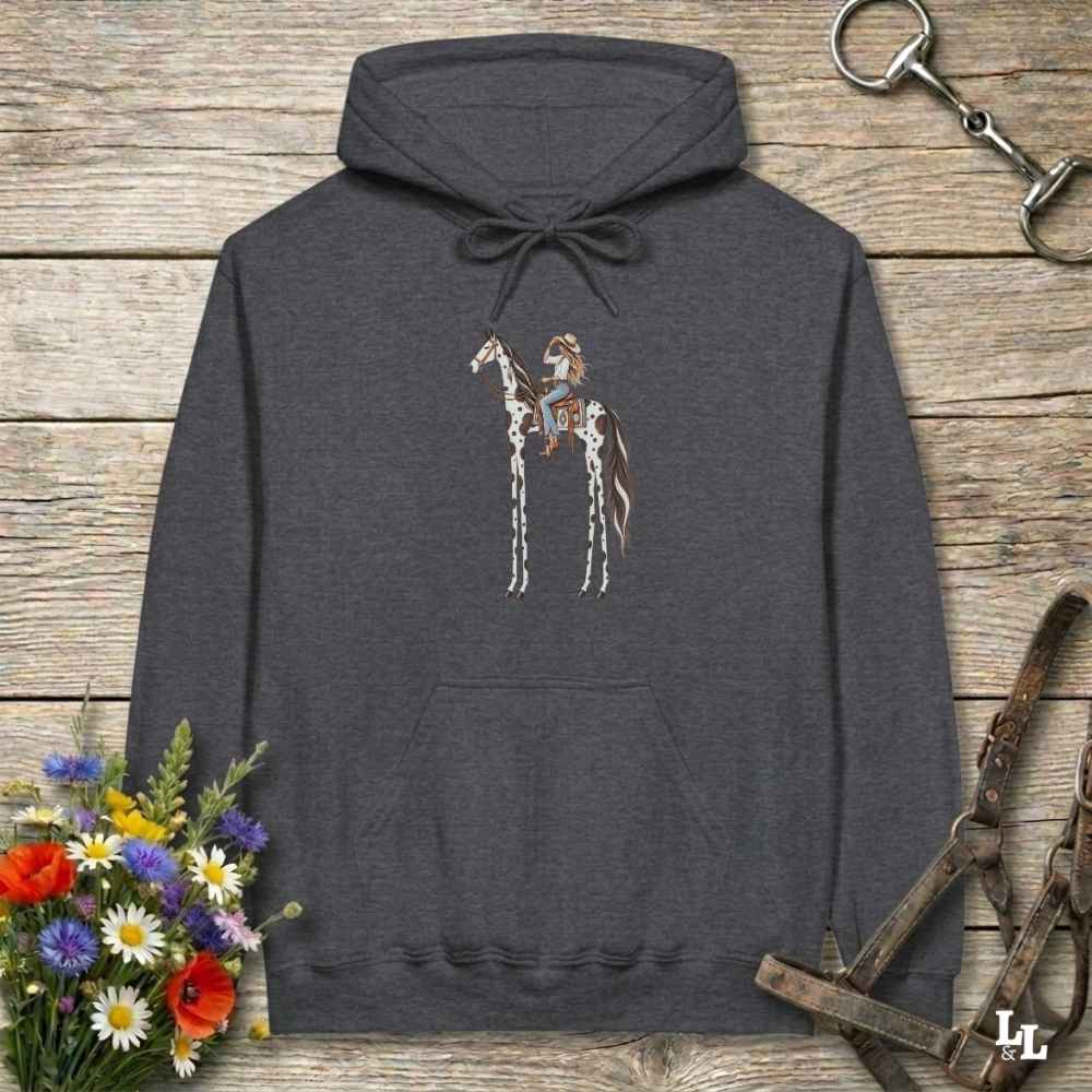 A dark heather gray hoodie laid flat on a rustic wooden surface, featuring a graphic of a tall horse with rider.. The hoodie is styled with equestrian-themed props including a halter, tack, and a small bouquet of wildflowers.